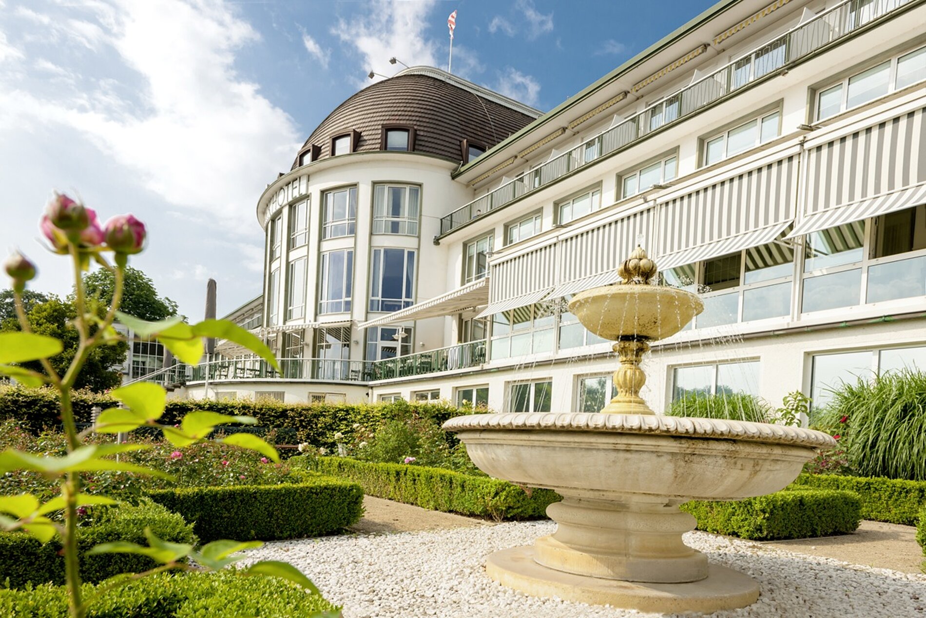 Helle Hotelfassade mit Brunnen und Rosengarten davor